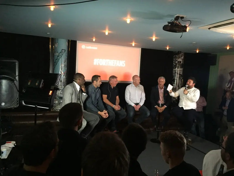 Grupa ludzi siedzących na scenie z prezenterem, przed publicznością.