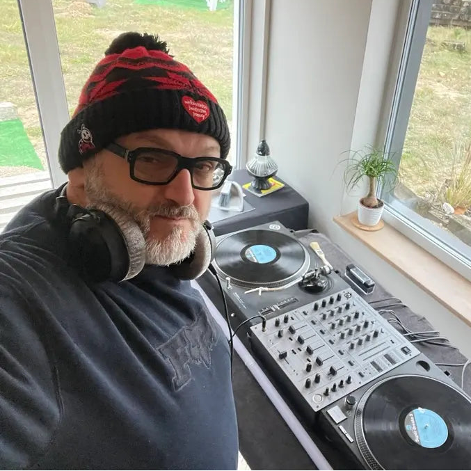 Osoba z słuchawkami i czapką beanie stojąca obok sprzętu DJ w pokoju z dużymi oknami, przez które widać widok na zewnątrz.