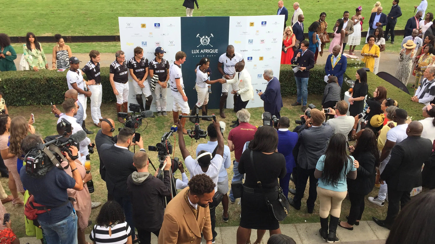 Wydarzenie z ludźmi na drewnianej platformie, otoczonymi tłumem i mediami, z polem trawiastym w tle.