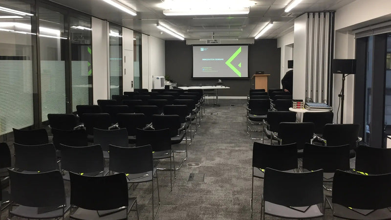 Sala konferencyjna z czarnymi krzesłami ustawionymi w rzędach, ekranem projekcyjnym i szklaną ścianą.